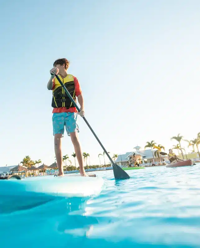 A person wearing a life vest stands on a paddleboard in a pool, holding a paddle, with buildings and palm trees visible in the background.