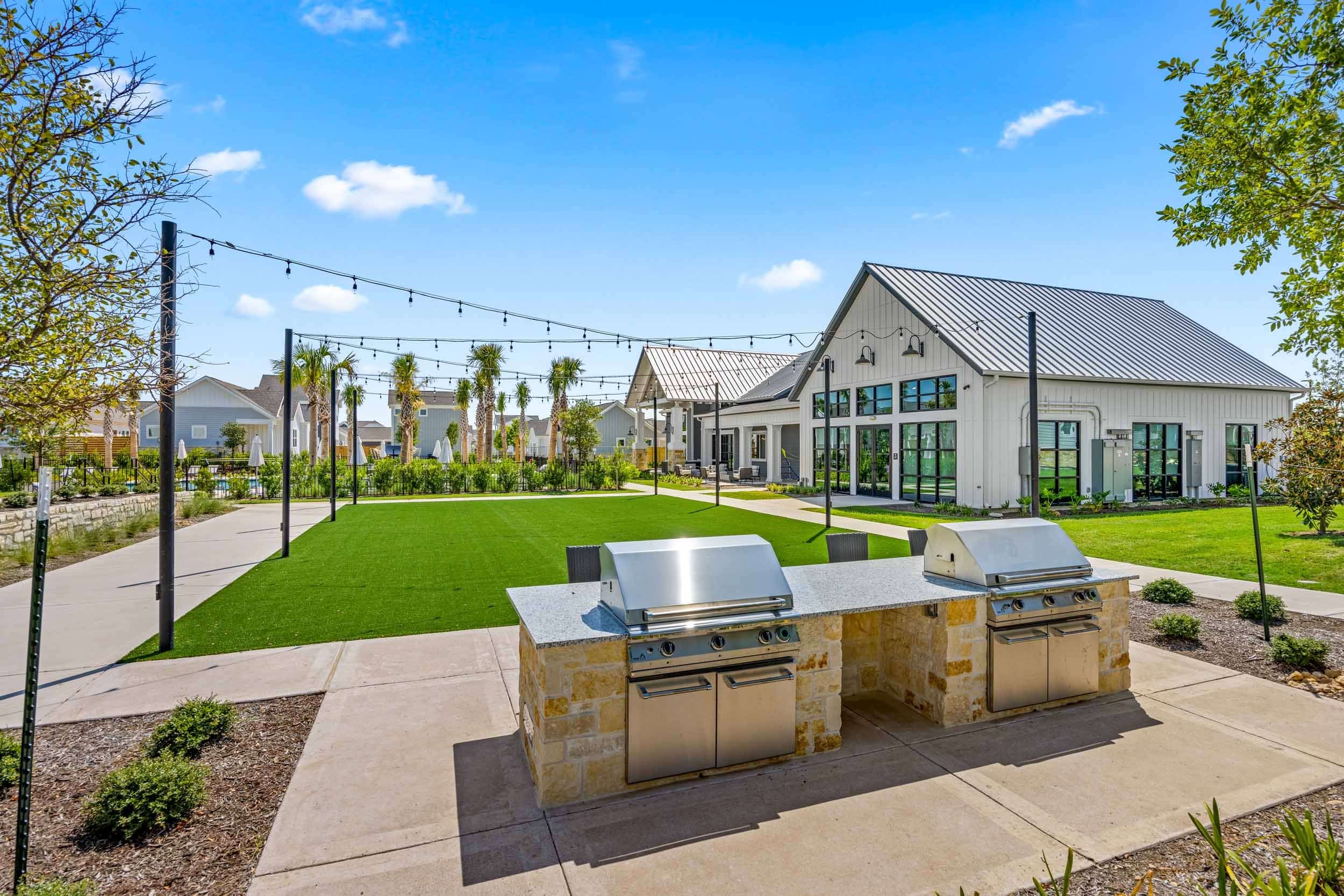 Outdoor community area with two stainless steel grills, string lights overhead, a large artificial turf lawn, and modern buildings in the background on a clear day.