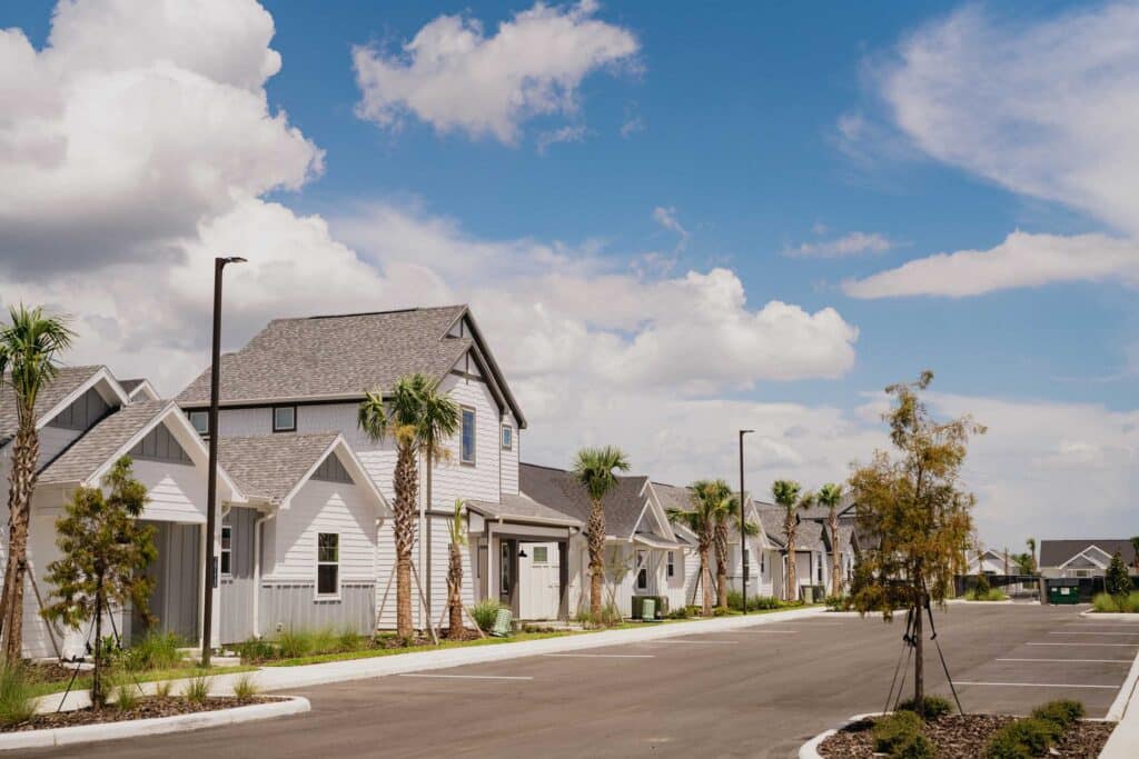 A row of modern suburban houses with driveways and palm trees lines a quiet street under a blue sky with scattered clouds.